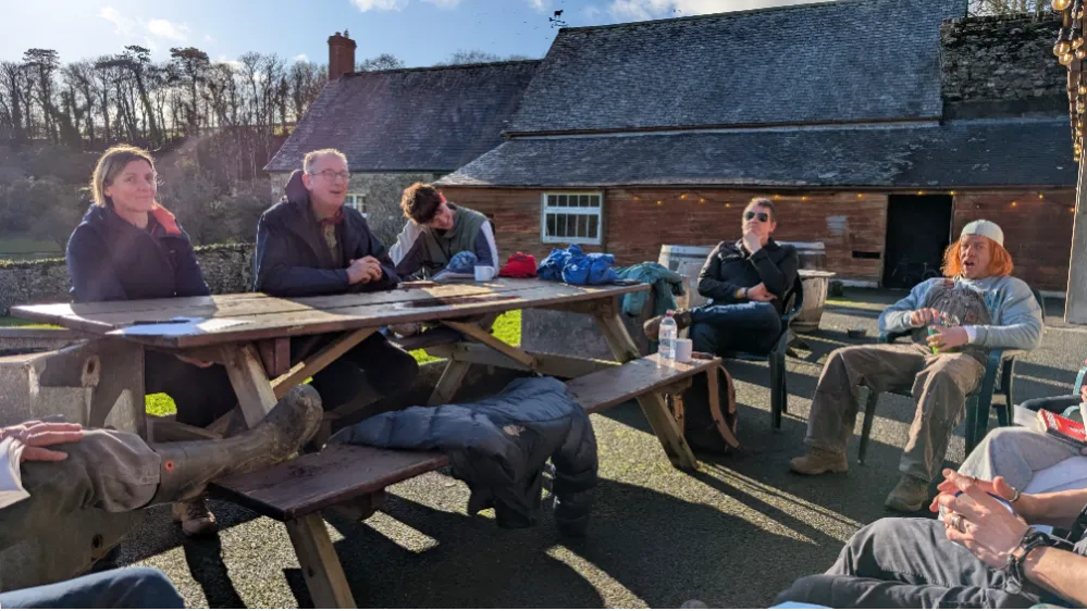 A group of people sitting around a picnic table and chairs outdoors enjoying a sunny day, with a rustic building in the background.
