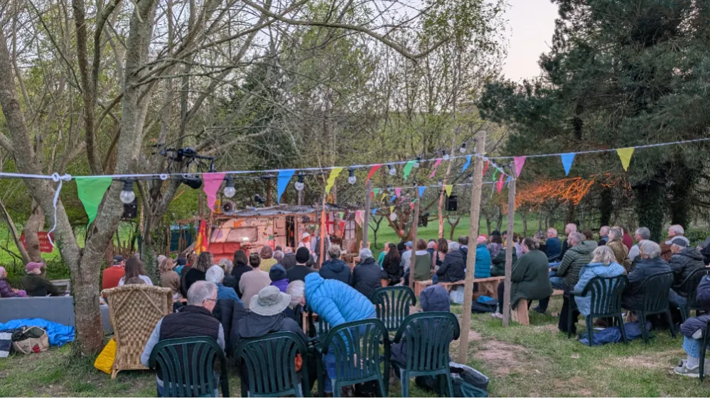 Outdoor gathering with people seated on chairs and benches watching a small stage, decorated with colorful bunting and surrounded by trees.