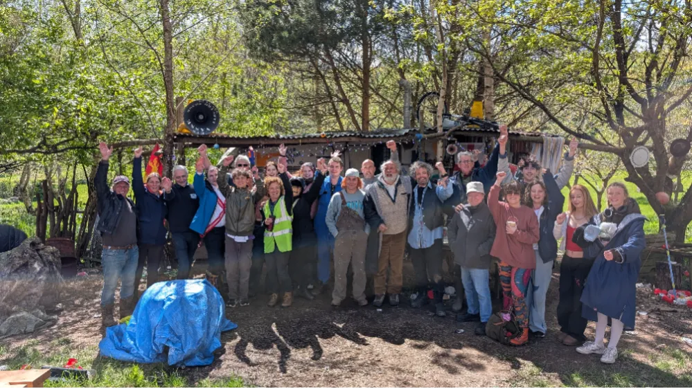 A large group of people posing together outdoors with raised arms in front of a rustic structure surrounded by trees.