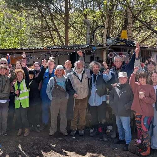 A large group of people posing together outdoors with raised arms in front of a rustic structure surrounded by trees.