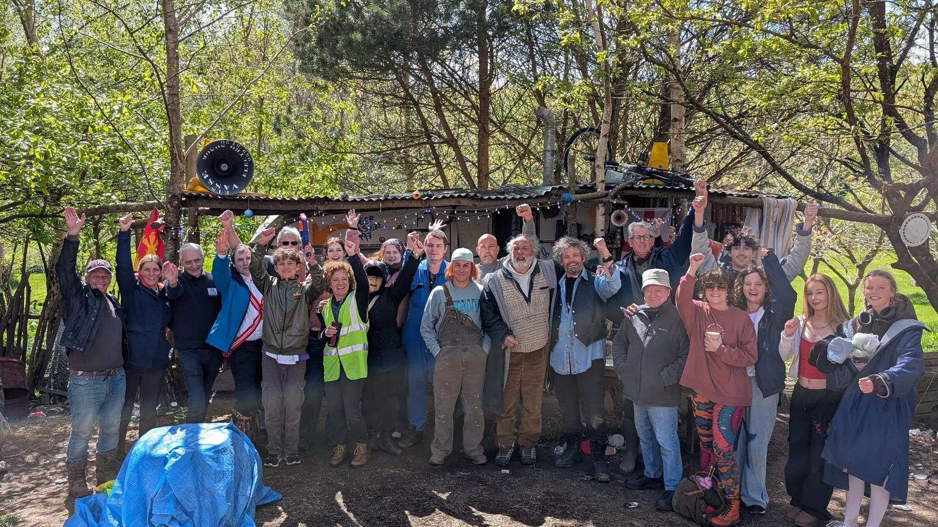 A large group of people posing together outdoors with raised arms in front of a rustic structure surrounded by trees.