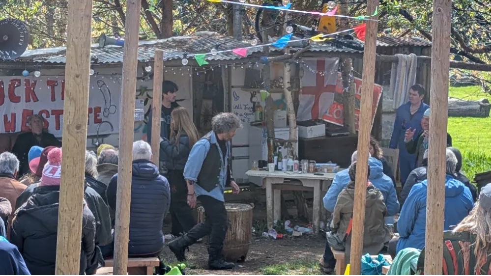 A group of people gathered outdoors in front of a rustic shed decorated with colorful bunting and flags, attending an event or meeting.