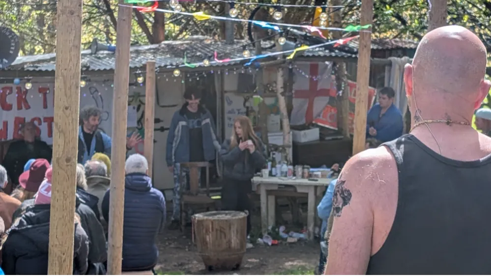 Outdoor gathering with people watching a group at a makeshift stage or bar, decorated with bunting and fairy lights.