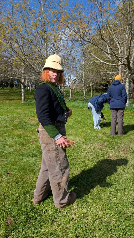 Person in a vintage-style jacket and helmet standing on grass, holding a piece of wood, with others working in the background.