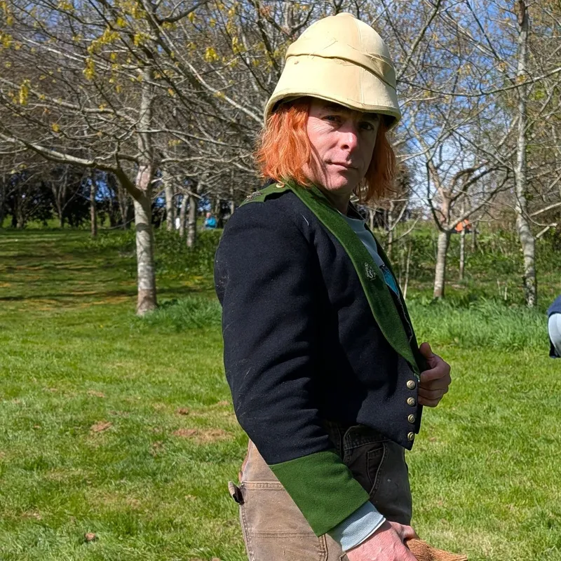 Person in a vintage-style jacket and helmet standing on grass, holding a piece of wood, with others working in the background.