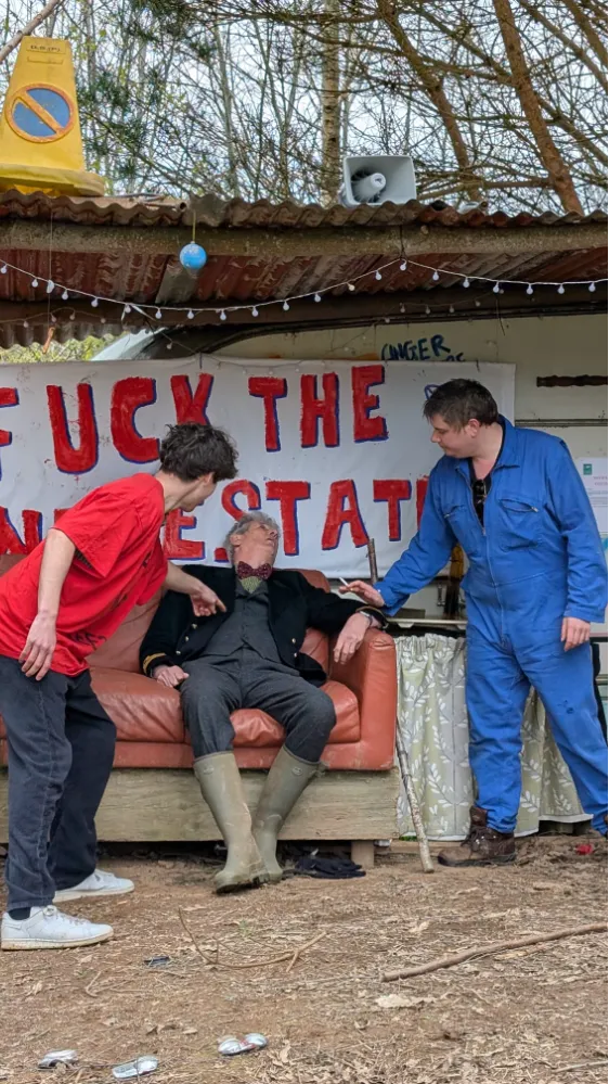 Three people in front of a handmade sign with a profane message, one sitting on a couch in costume, two standing and interacting with him.