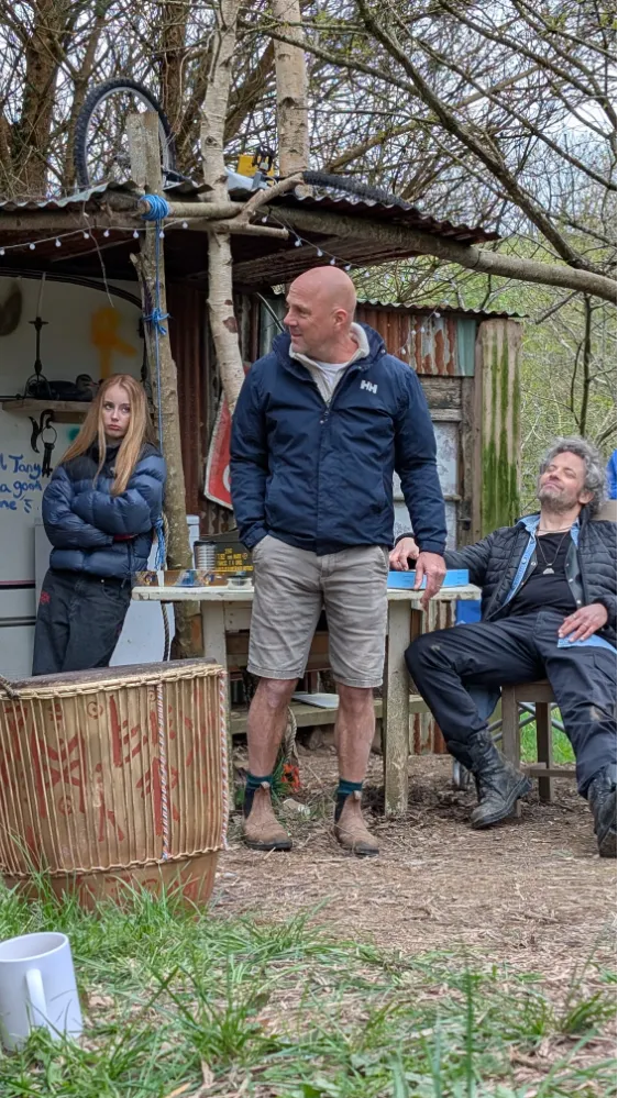 Three people outdoors near a rustic shed, one standing, one leaning on the table, and one sitting in a chair