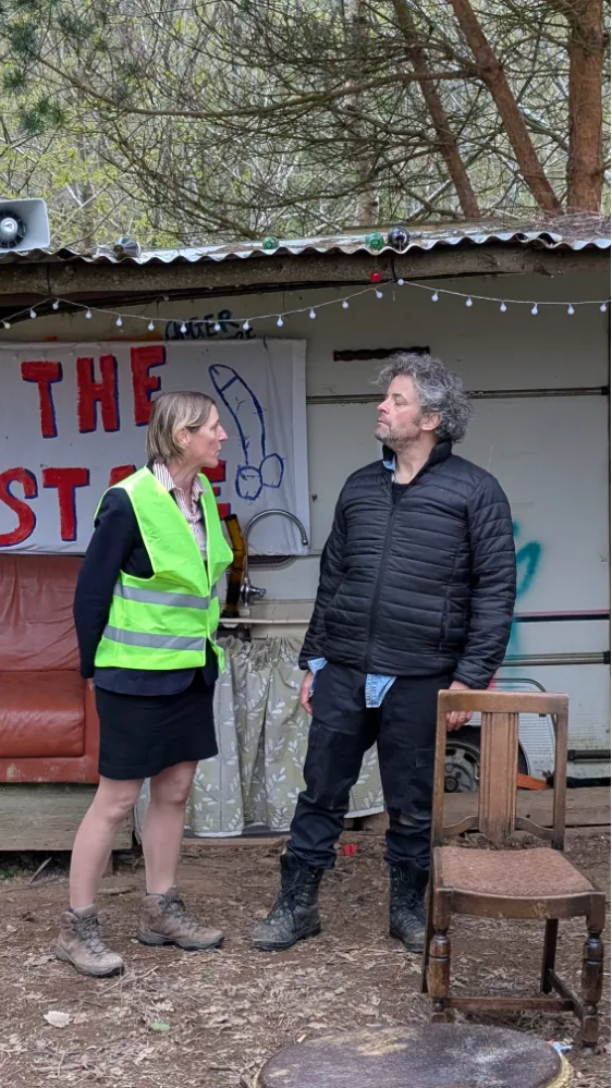 Two people standing and talking in front of a makeshift shelter with a banner and forest background.