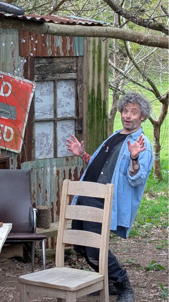 Person with curly hair and denim shirt playfully leaning back near rustic shed.
