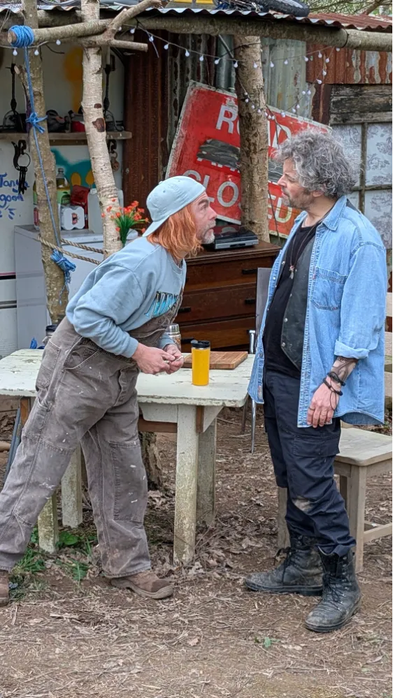 Two men having an intense conversation outside in a rustic, makeshift setting with trees, a table, old furniture and a 'Road Closed' sign.