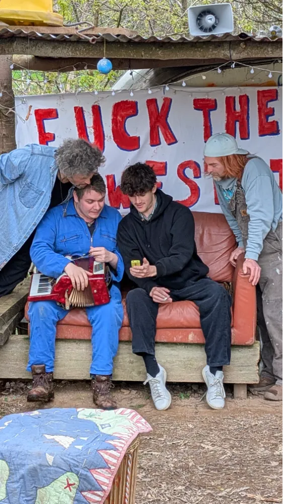 Four people gathered outside on a couch, one holding an accordion, another showing a phone, with a painted banner in the background containing explicit language.