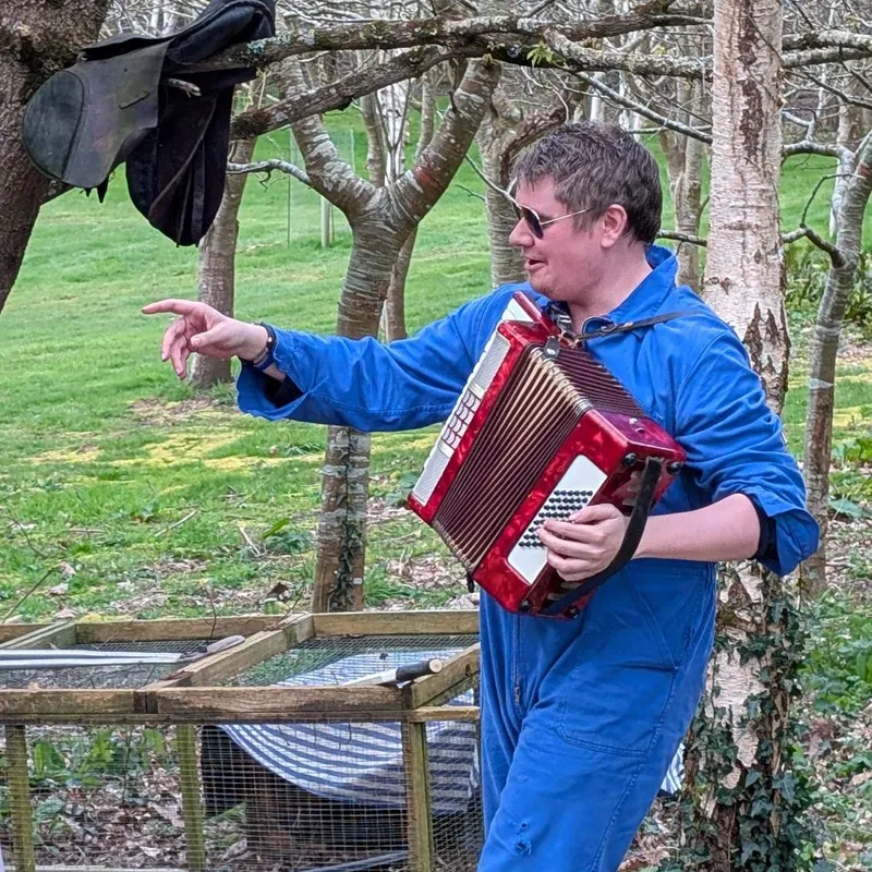 Person in blue jumpsuit playing a red accordion outdoors near trees