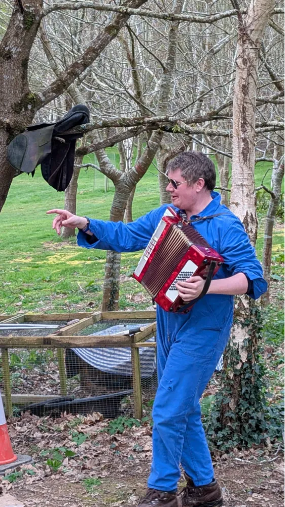 Person in blue jumpsuit playing a red accordion outdoors near trees