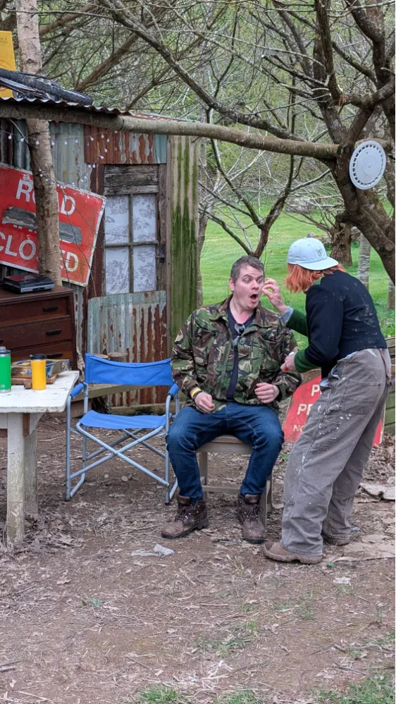 Person sitting on a chair with another person standing in front, appearing to inspect or touch the other's face, outdoors near a rustic shed
