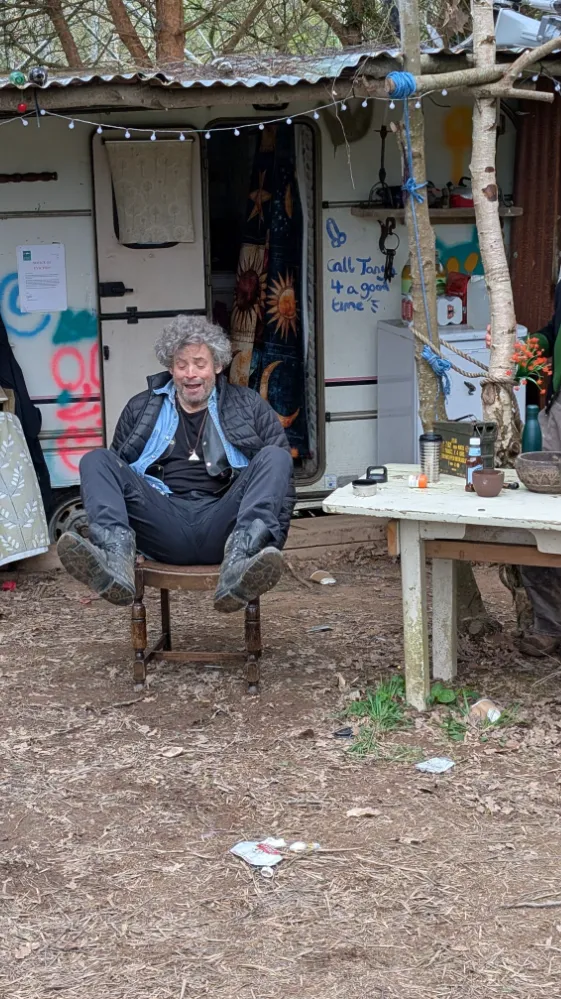 A man with curly gray hair sits on a wooden chair outside a caravan, appearing to laugh or shout, with legs raised. A table and some trees are beside him.