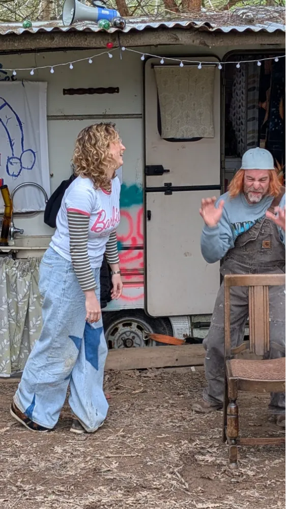 Two people interacting playfully outside a rustic caravan, with one person standing and smiling and the other seated and gesturing with their hands.