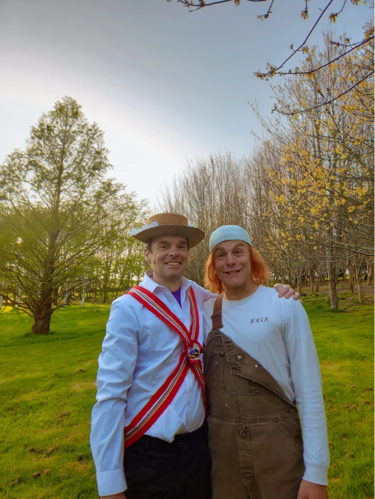 Two people posing in a park, one wearing a straw hat and red sash, the other in a bandana and brown overalls.