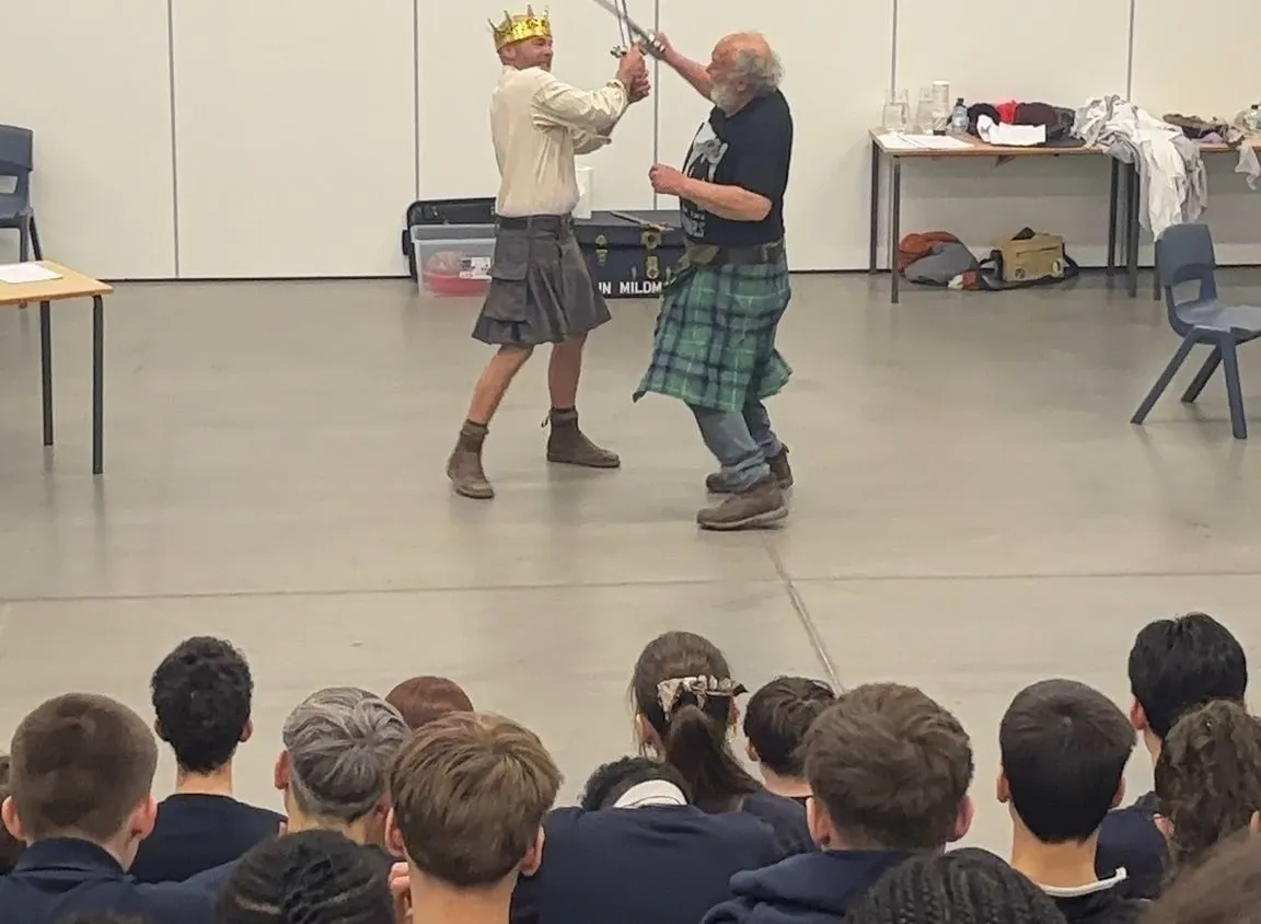 Two men in kilts perform a staged sword fight in front of a seated audience in a hall, with one wearing a gold crown.
