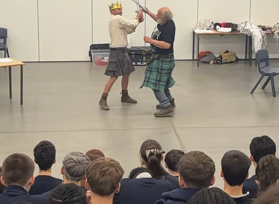 Two men in kilts perform a staged sword fight in front of a seated audience in a hall, with one wearing a gold crown.