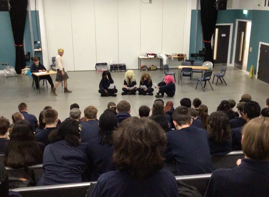 Students perform a drama scene in a school hall while an audience watches from tiered seating.