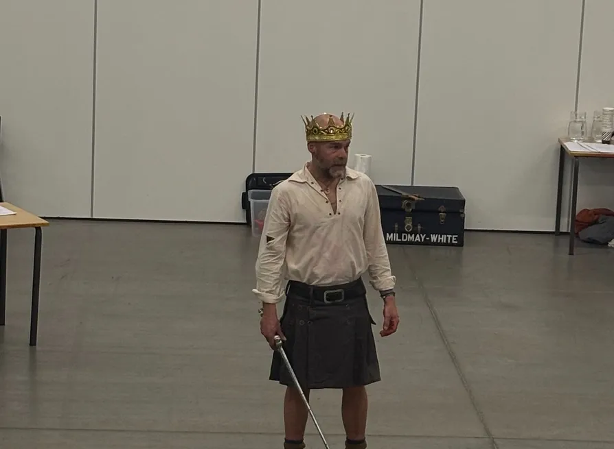 Actor in a crown and kilt holding a sword during a rehearsal or performance in a large hall