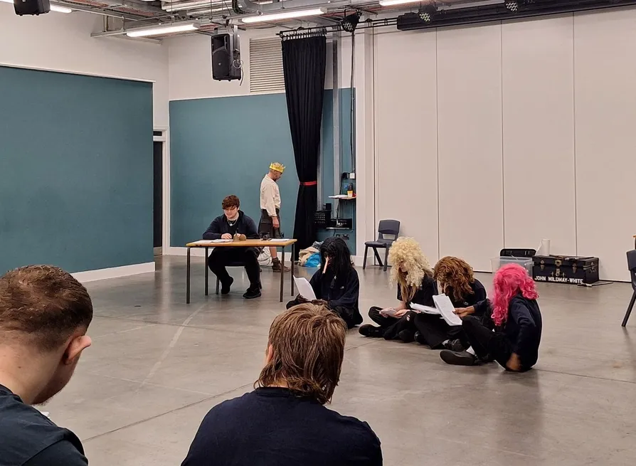Students rehearsing a scene in a drama studio, with performers seated on the floor reading scripts and one person wearing a paper crown near a table.