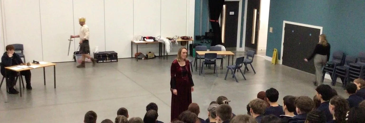 Students watch a live performance in a school theatre, with actors on stage and simple props arranged around the space.
