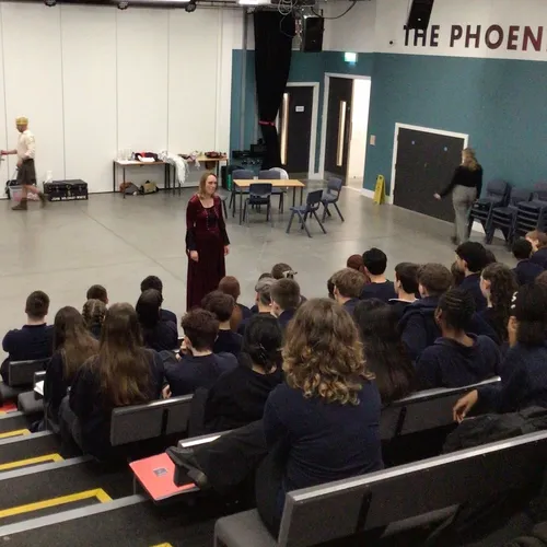 Students watch a live performance in a school theatre, with actors on stage and simple props arranged around the space.