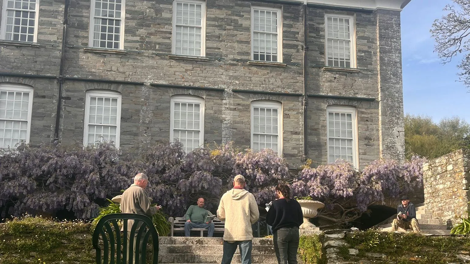 Cast members rehearse The Crucible on the lawn in front of a large stone house with blooming wisteria.