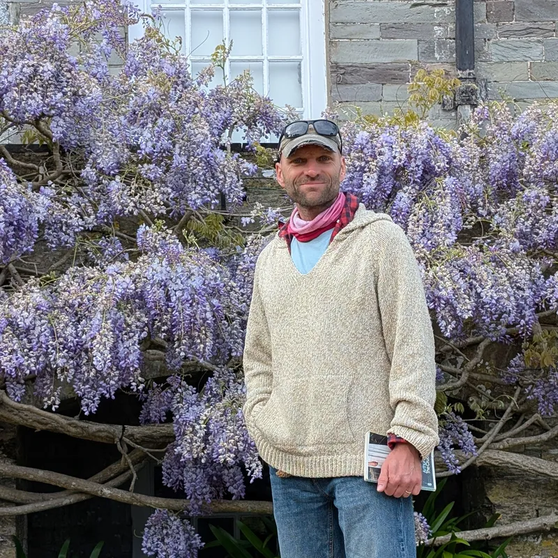 A man standing in front of blooming purple wisteria against a stone house wall, holding a book.