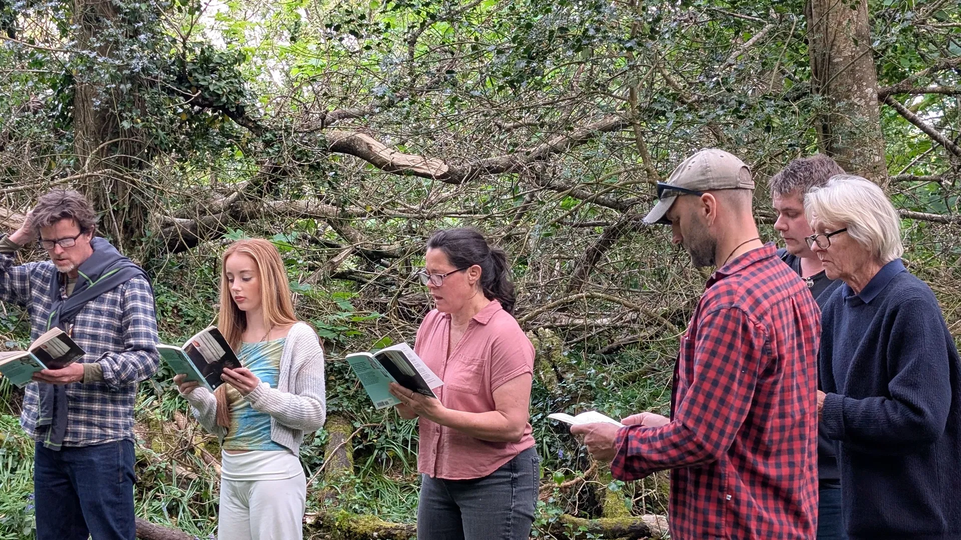 Cast members rehearse The Crucible outdoors in a wooded area, standing in a line and reading from scripts.