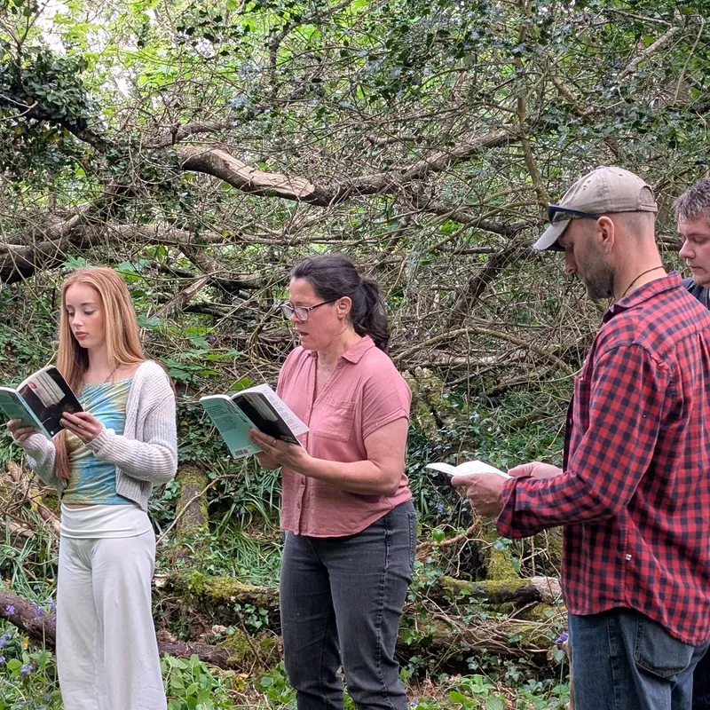 Cast members rehearse The Crucible outdoors in a wooded area, standing in a line and reading from scripts.