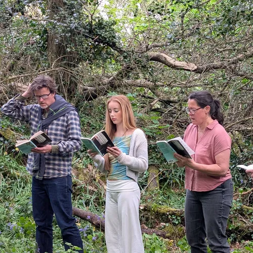 Cast members rehearse The Crucible outdoors in a wooded area, standing in a line and reading from scripts.