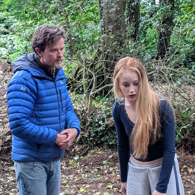 Two performers rehearse a scene from The Crucible in a wooded outdoor setting, one in a blue jacket and the other in grey trousers and a black top.