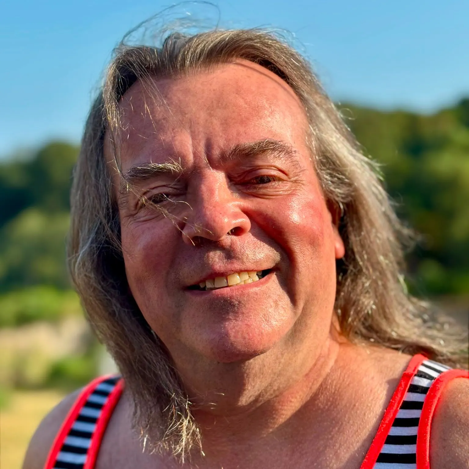 Gregory Max Paul smiling outdoors in bright sunshine, wearing a striped sleeveless top with red trim.