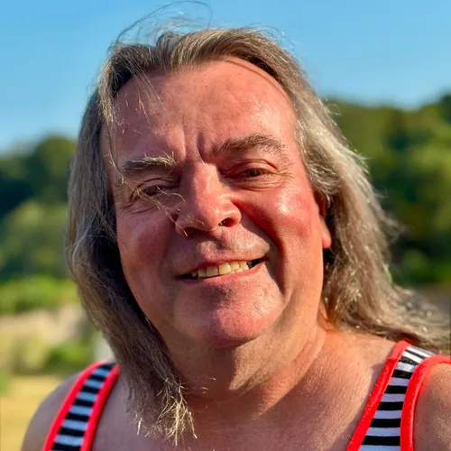 Gregory Max Paul smiling outdoors in bright sunshine, wearing a striped sleeveless top with red trim.