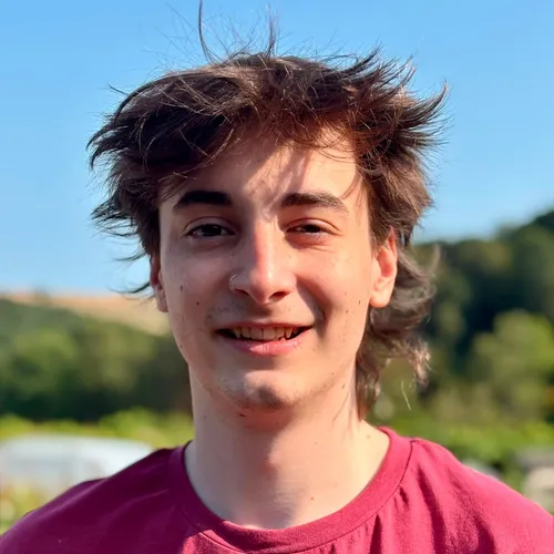Portrait of Mercutio Ethan Payne outdoors in a burgundy T-shirt, smiling in sunlight with trees and blue sky behind him.