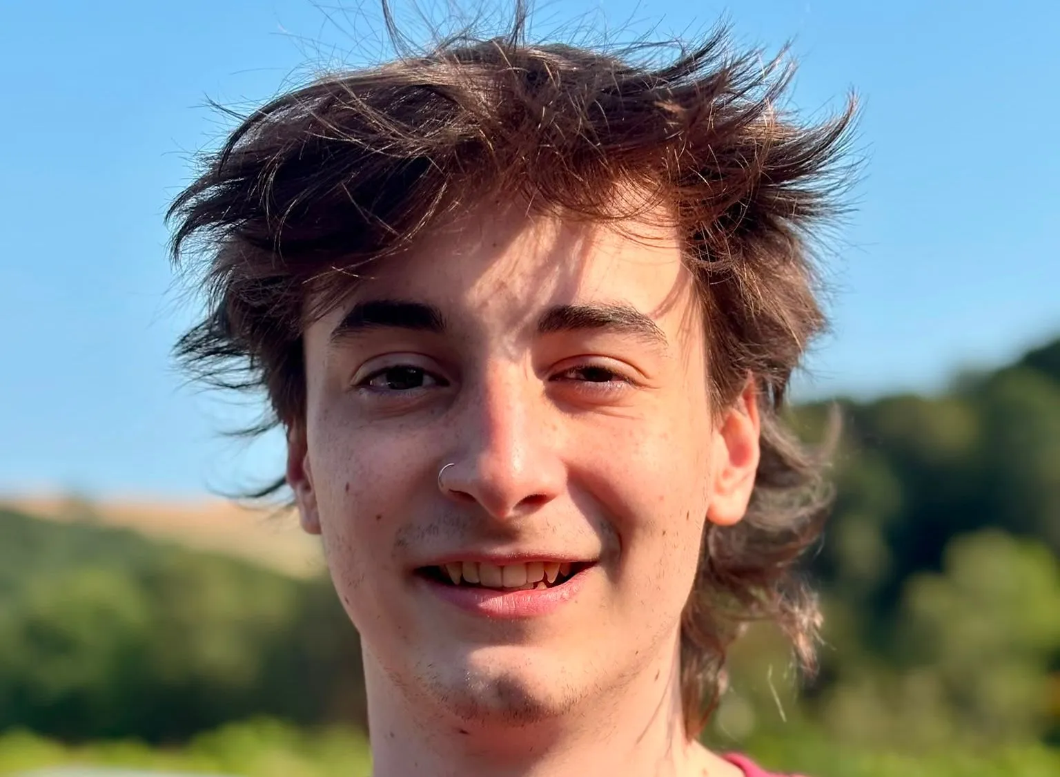 Portrait of Mercutio Ethan Payne outdoors in a burgundy T-shirt, smiling in sunlight with trees and blue sky behind him.