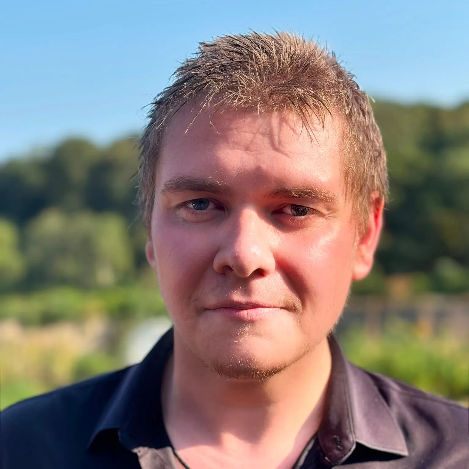 Portrait of John Currall outdoors in warm sunlight, wearing a black shirt with a blurred green landscape behind him.