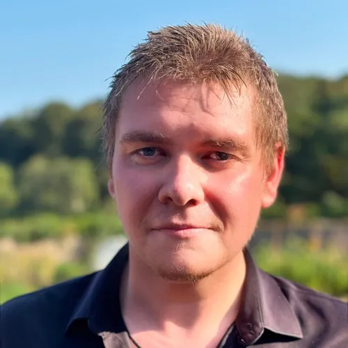Portrait of John Currall outdoors in warm sunlight, wearing a black shirt with a blurred green landscape behind him.