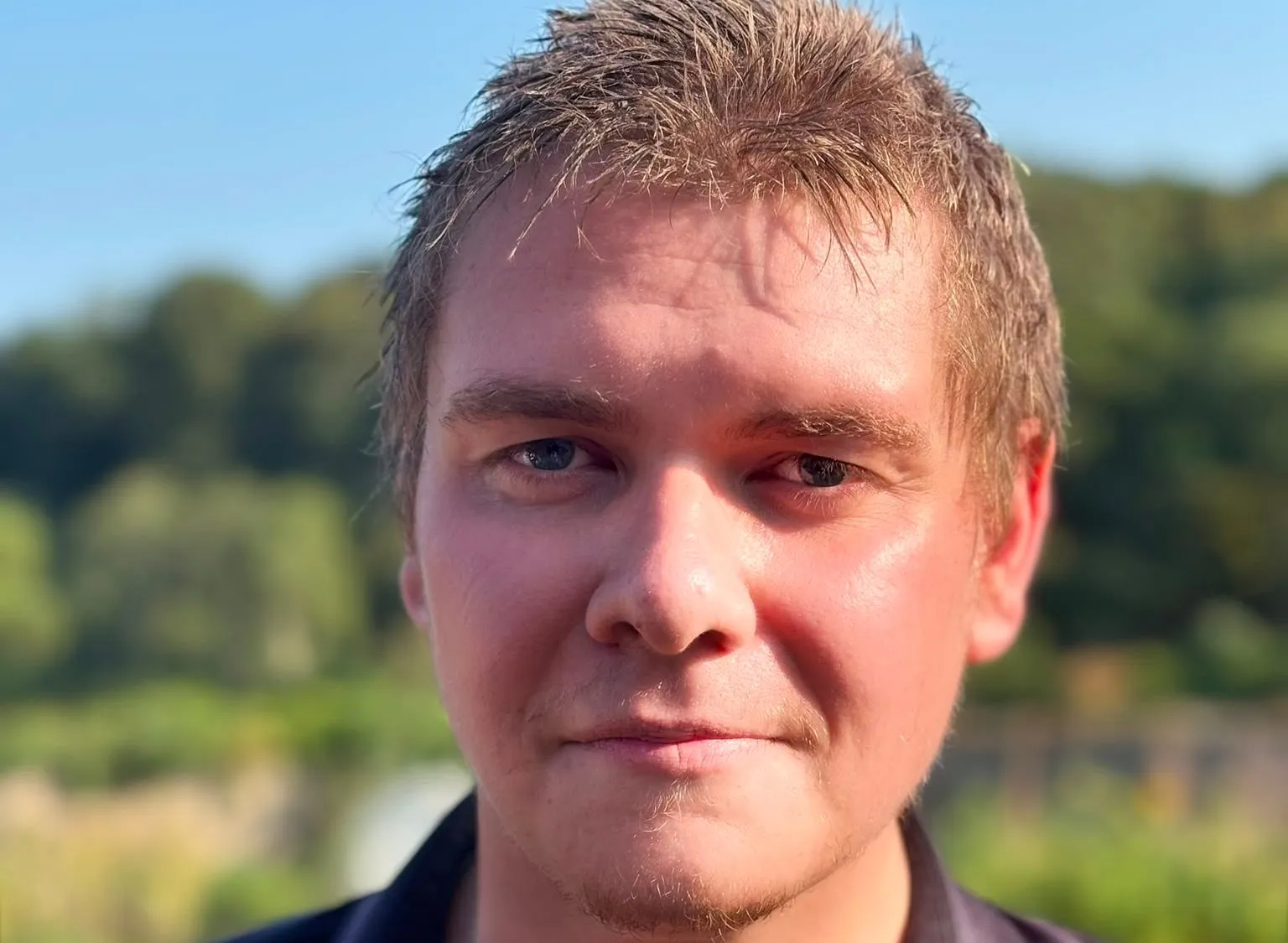 Portrait of John Currall outdoors in warm sunlight, wearing a black shirt with a blurred green landscape behind him.
