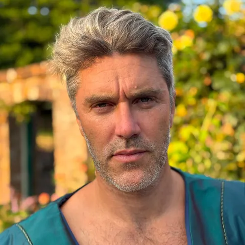 Portrait of Tybalt Howie Quinlivan outdoors in period-style clothing, with silver hair and a blurred garden background.