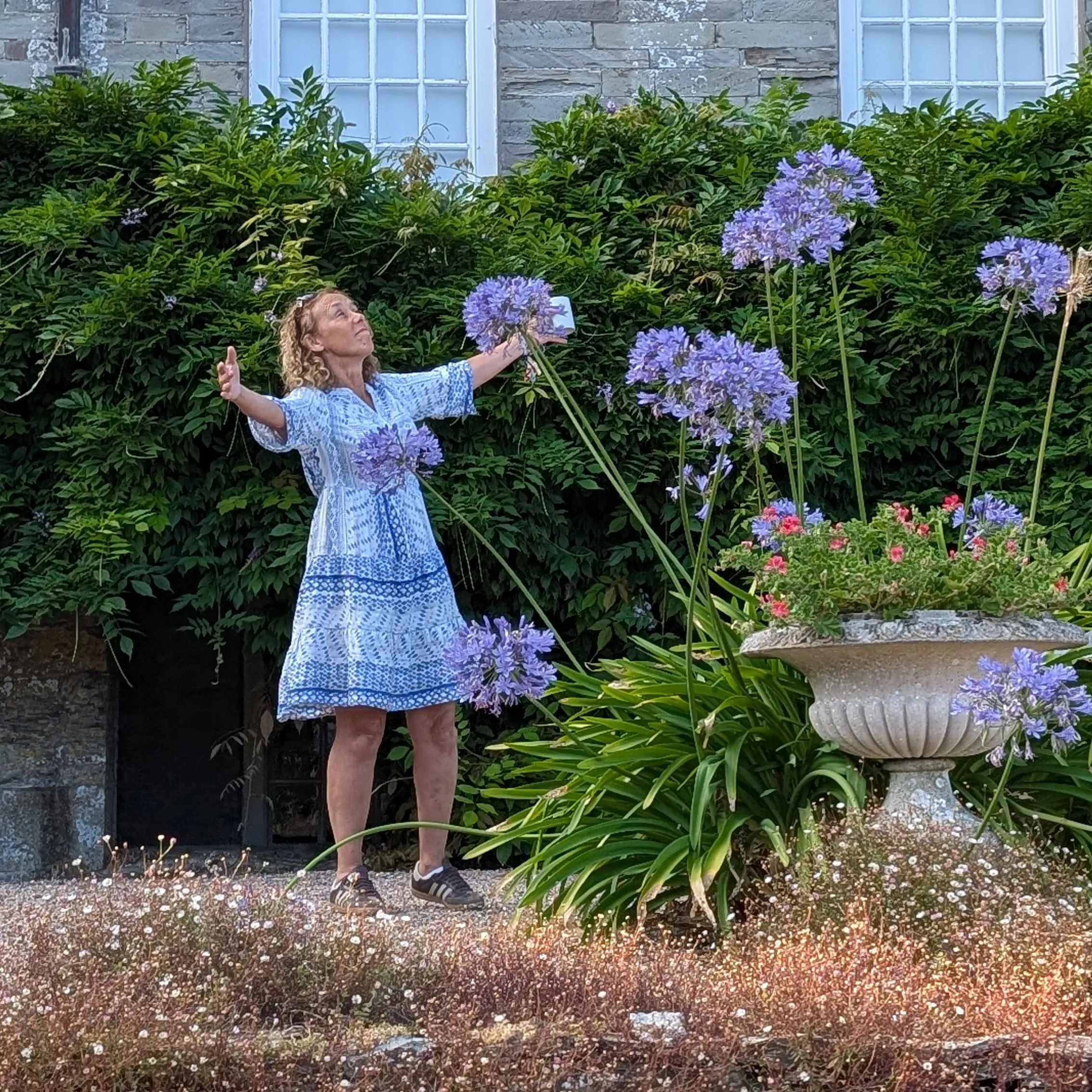 Woman rehearsing with outstretched arms in a flower-filled garden beside an old stone building
