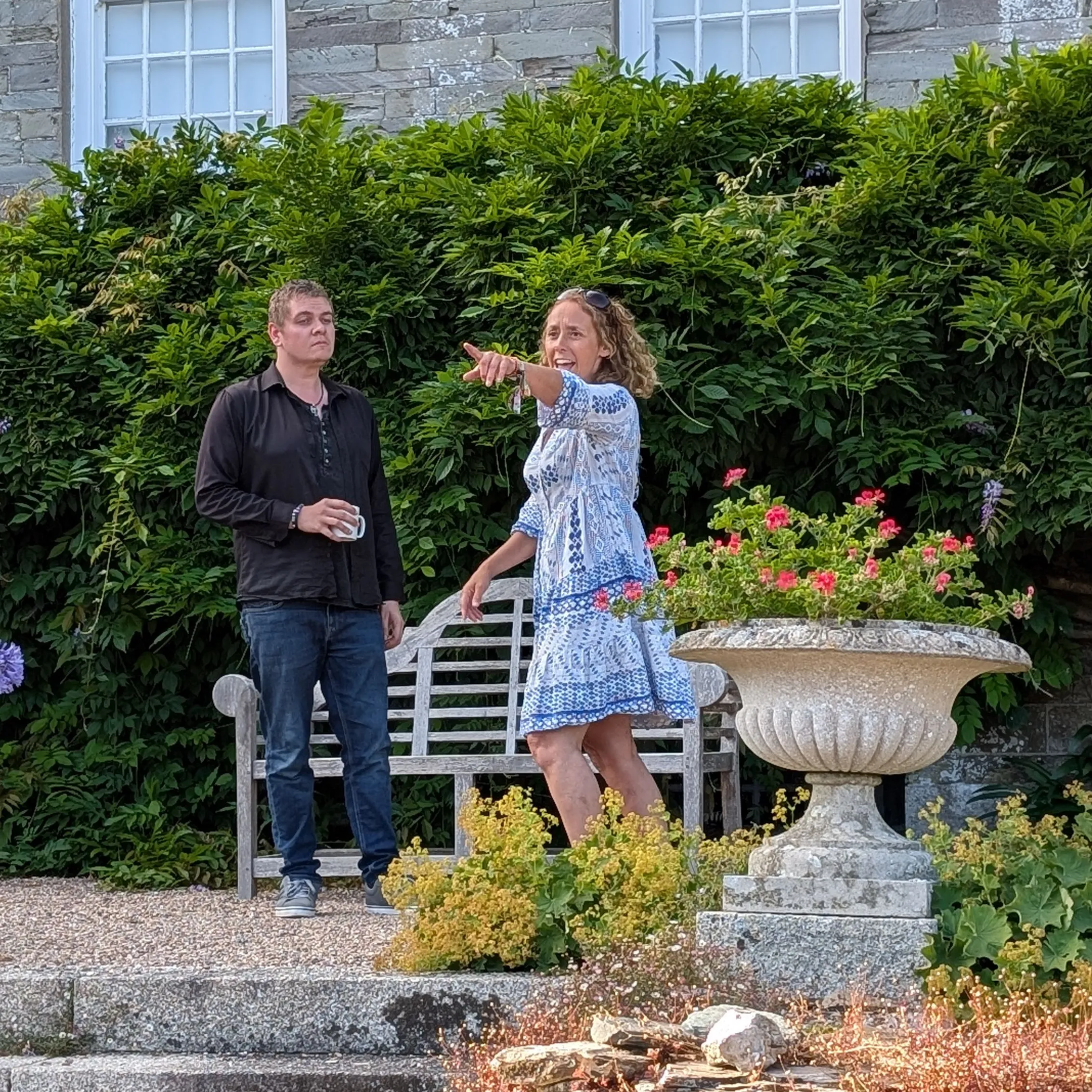 Two performers rehearsing in a historic garden, with one woman pointing beside a bench and stone urn.