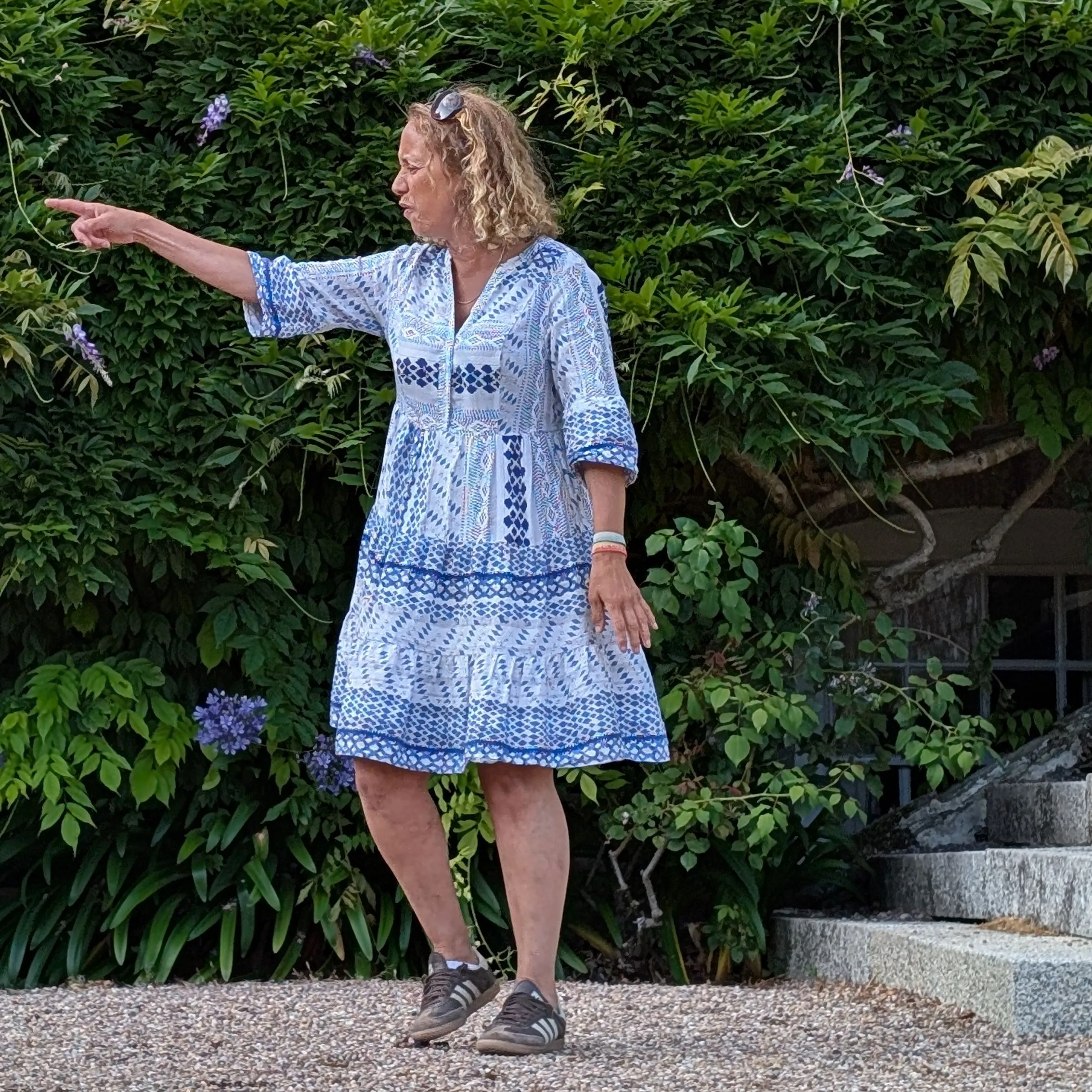 Woman in a blue patterned dress pointing beside a leafy garden and brick building