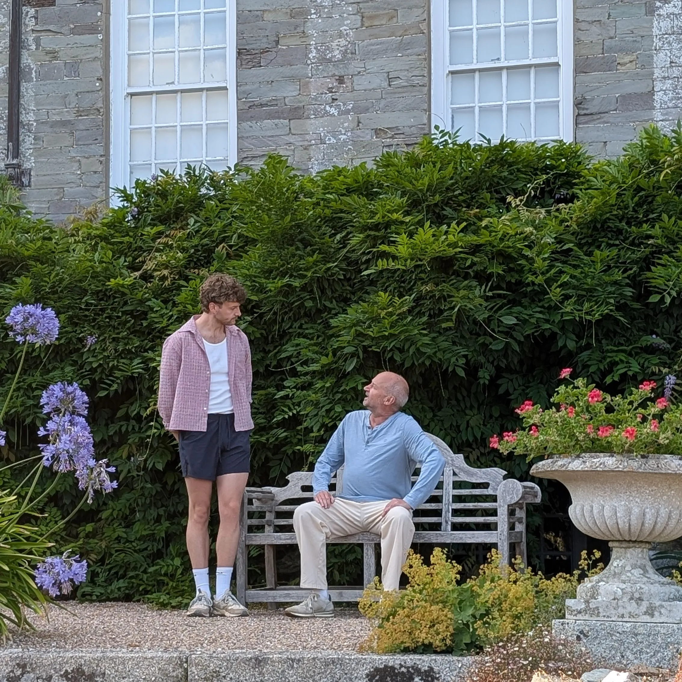 Two men rehearsing a scene beside a bench in the garden of a historic stone house.