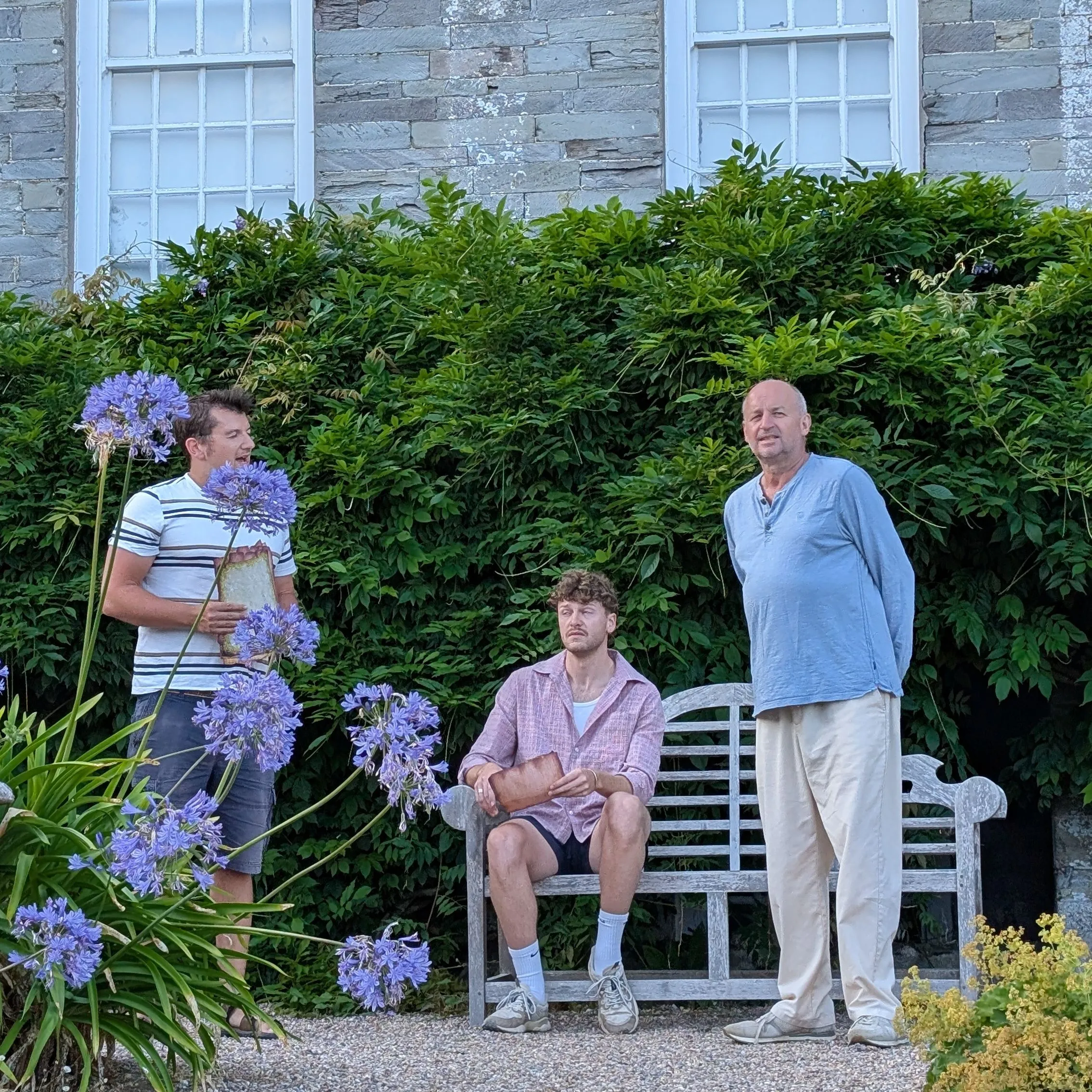 Three men rehearsing outdoors in a garden beside a stone building, with one seated on a bench holding a script.