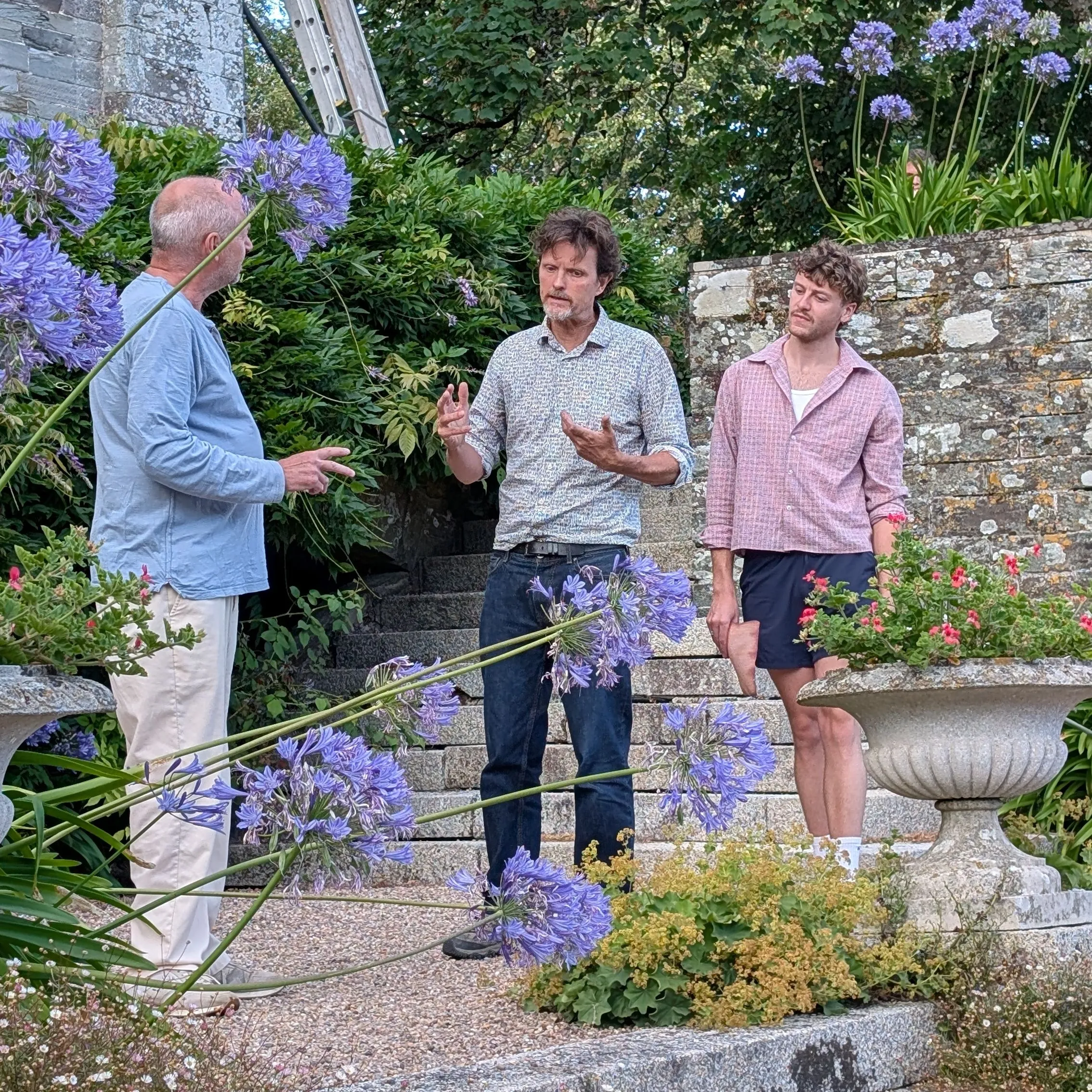 Three men rehearsing a scene from The Crucible in a flower-filled garden beside a historic stone building.