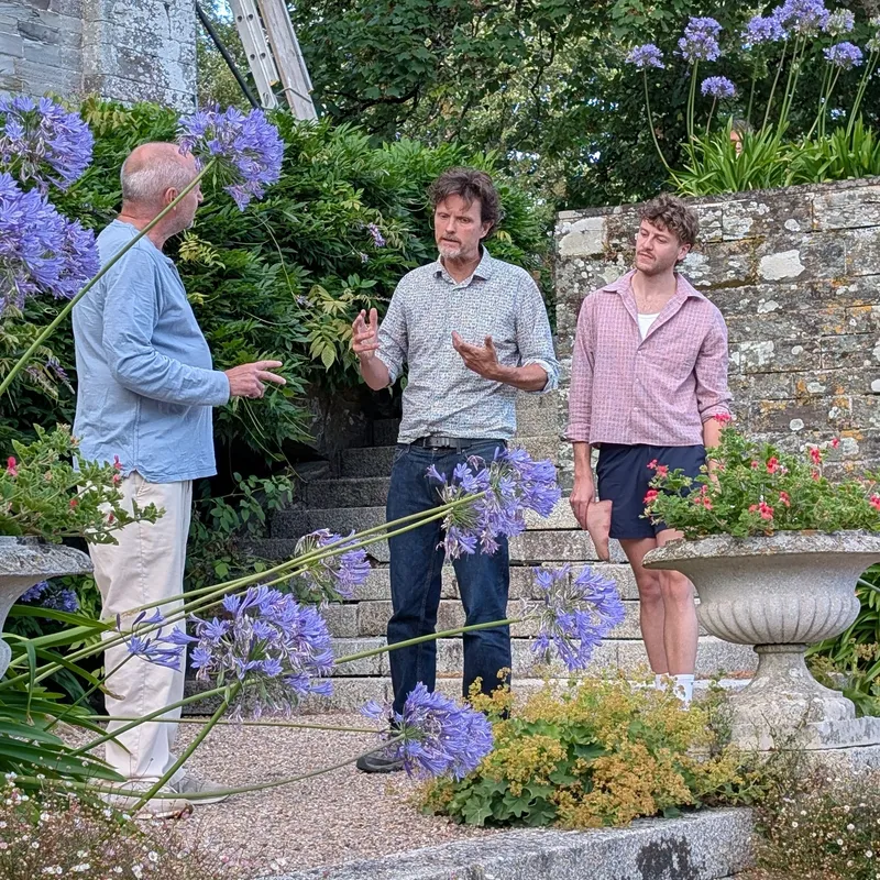 Three men rehearsing a scene from The Crucible in a flower-filled garden beside a historic stone building.