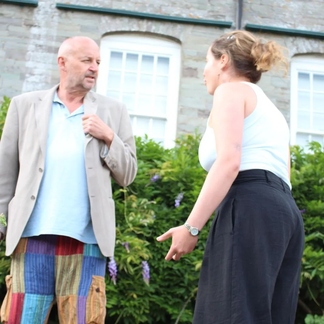 Two actors rehearsing an outdoor scene in front of a stone building.
